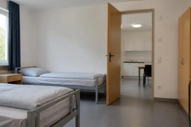Άδεια κρεβάτια σε δωμάτιο συλλογικής φιλοξενίας με κοινόχρηστο χώρο στο βάθος.