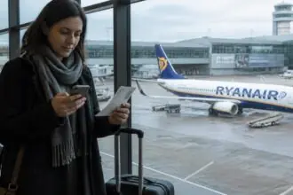 Ταξιδιώτης ελέγχει το κινητό του σε αίθουσα αναχωρήσεων, με αεροσκάφος της Ryanair στο βάθος.