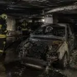 Καμένο αυτοκίνητο σε υπόγειο πάρκινγκ με πυροσβέστες να εξετάζουν τον χώρο.