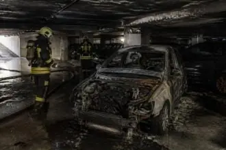 Καμένο αυτοκίνητο σε υπόγειο πάρκινγκ με πυροσβέστες να εξετάζουν τον χώρο.