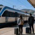 Symbolisches Bild eines stehenden Personenzugs an einem griechischen Bahnhof mit wartenden Reisenden