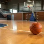 Ein Basketball liegt auf dem Parkett einer leeren Sporthalle, im Hintergrund ist ein Basketballkorb zu sehen.