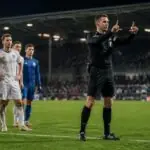 Διαιτητής κάνει το σήμα του VAR δίπλα σε ποδοσφαιριστές που περιμένουν την απόφασή του σε γήπεδο.