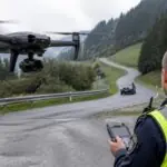 Drone με κάμερα αιωρείται κοντά σε ορεινό δρόμο της Ελβετίας, ενώ χειριστής κρατά τηλεχειριστήριο.