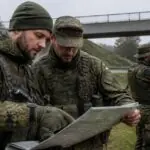 Έφεδροι της Bundeswehr με στρατιωτικές στολές συμμετέχουν σε υπαίθρια άσκηση εκπαίδευσης κοντά σε γέφυρα.