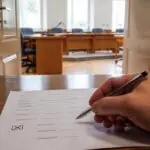 Χέρι κρατά στυλό πάνω από έντυπο επιλογής, με αίθουσα δημοτικού συμβουλίου στο βάθος.