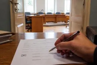 Χέρι κρατά στυλό πάνω από έντυπο επιλογής, με αίθουσα δημοτικού συμβουλίου στο βάθος.