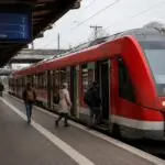 Επιβάτες περπατούν σε αποβάθρα γερμανικού σταθμού δίπλα σε κόκκινο περιφερειακό τρένο.