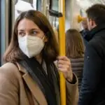 Επιβάτιδα με λευκή μάσκα FFP2 κρατά χειρολαβή μέσα σε αστικό μέσο μεταφοράς με άλλους επιβάτες.