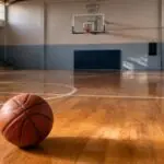Ein Basketball liegt auf dem Parkett einer leeren Sporthalle mit Basketballkorb im Hintergrund.