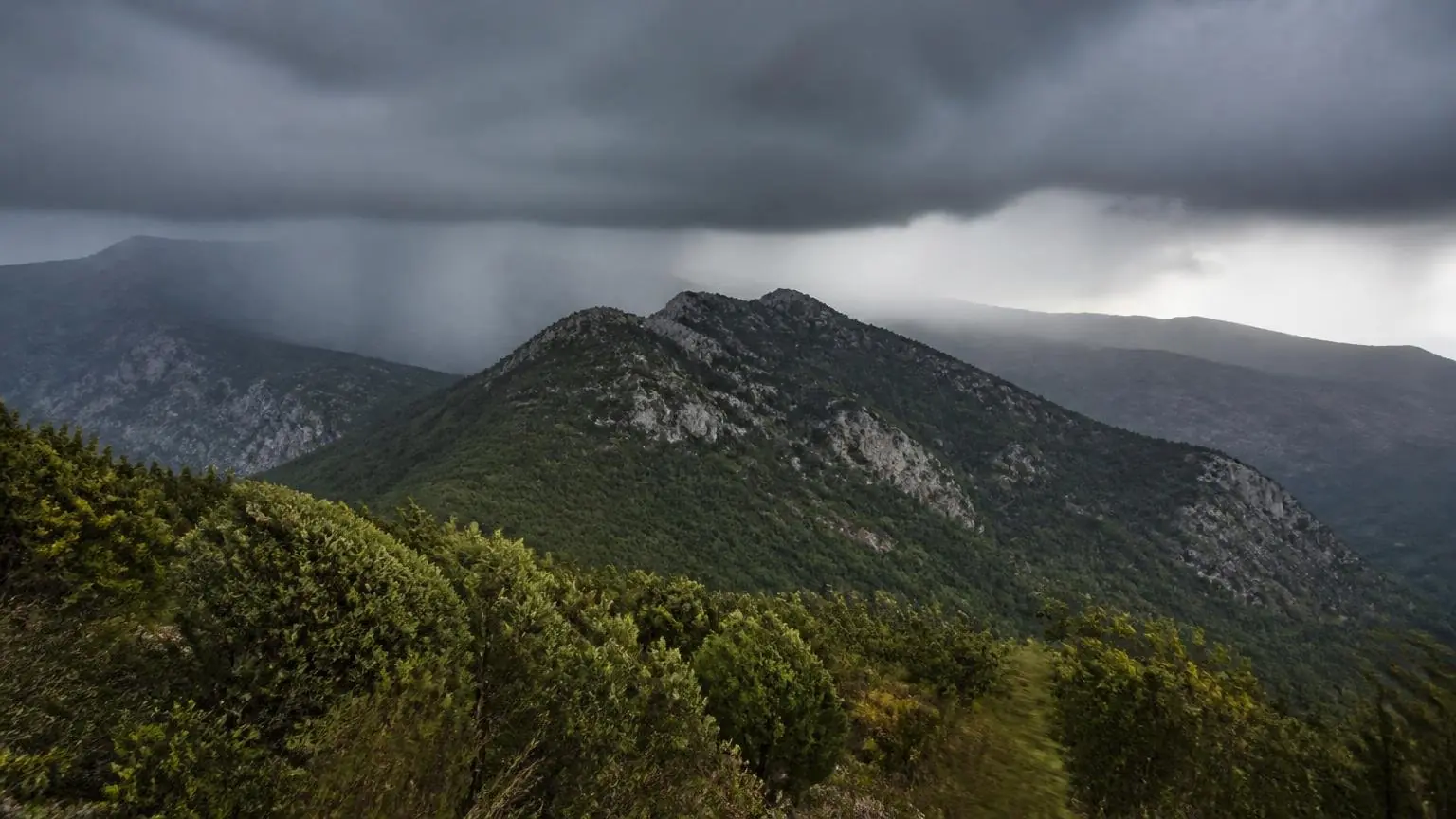 Dunkle Regenwolken über bewaldeten Bergen in einer griechischen Landschaft.
