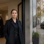 Νεαρή επαγγελματίας έξω από γραφείο τελετών στη Γερμανία, με διακριτικό όχημα κηδειών στο βάθος.
