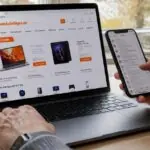 Άτομο ελέγχει το κινητό του δίπλα σε ανοιχτό φορητό υπολογιστή με ηλεκτρονικό κατάστημα στην οθόνη.