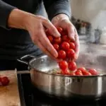 Χέρια ρίχνουν κόκκινα ντοματίνια σε κατσαρόλα σε οικιακή κουζίνα για μαγείρεμα.