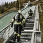 Πυροσβέστες με πλήρη εξοπλισμό ανεβαίνουν υπαίθρια σκάλα δίπλα σε εξέδρα άλματος σκι.
