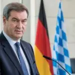 Συμβολικό πορτρέτο του Markus Söder σε γερμανικό πολιτικό περιβάλλον με σημαίες στο βάθος