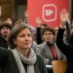 Συμμετέχοντες σε συμβολική πολιτική συνέλευση στην Ελβετία σηκώνουν τα χέρια για ψηφοφορία, με κόκκινο φόντο.