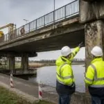Μηχανικοί επιθεωρούν παλαιότερη γέφυρα πάνω από υδάτινη οδό σε συμβολική σκηνή συντήρησης υποδομών στην Ολλανδία.