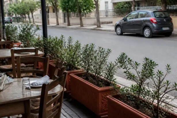 Leere Tische und Stühle eines griechischen Lokals stehen hinter Pflanzkübeln an einer Straße; im Hintergrund ist ein parkendes Auto zu sehen.