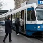 Επιβάτες κατευθύνονται προς μπλε τραμ σε στάση των δημόσιων συγκοινωνιών της Ζυρίχης.