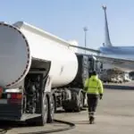 Βυτιοφόρο ανεφοδιασμού καυσίμων σε χώρο αεροδρομίου, με επιβατικό αεροπλάνο στο βάθος.